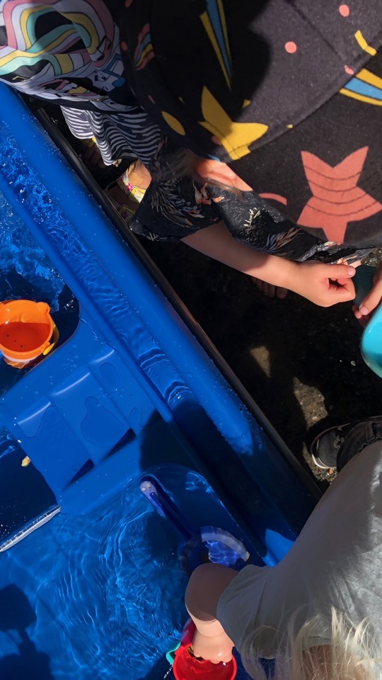 Ett barn med keps leker med vatten och leksaker i en blå vattenlek på sommaren.