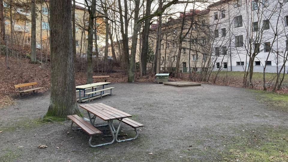 Lekplats med sandlåda och parkbänkar, omgiven av träd och med flerfamiljshus i bakgrunden. 