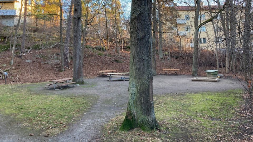 Lekplats med sandlåda och parkbänkar, omgiven av träd och med flerfamiljshus i bakgrunden. 