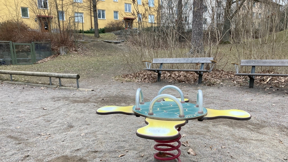 Yta med sand. I förgrunden en gungleksak. I bakgrunden en stock att gå balansgång på, bänkar, buskar och flerfamiljshus. 
