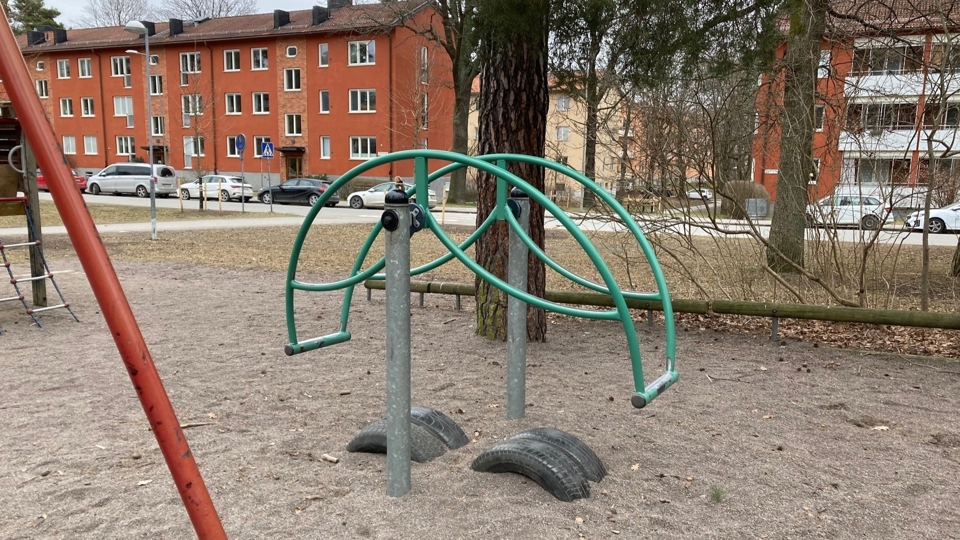 Yta med sand. I förgrunden en vippgunga. I bakgrunden träd, en gata med parkerade bilar och flerfamiljhus, foto.