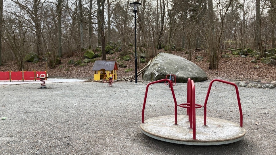 En lekkarusell på en lekplats med gungdjur och lekhus. Bredvid lekplatsen finns en skog med många stora stenar.