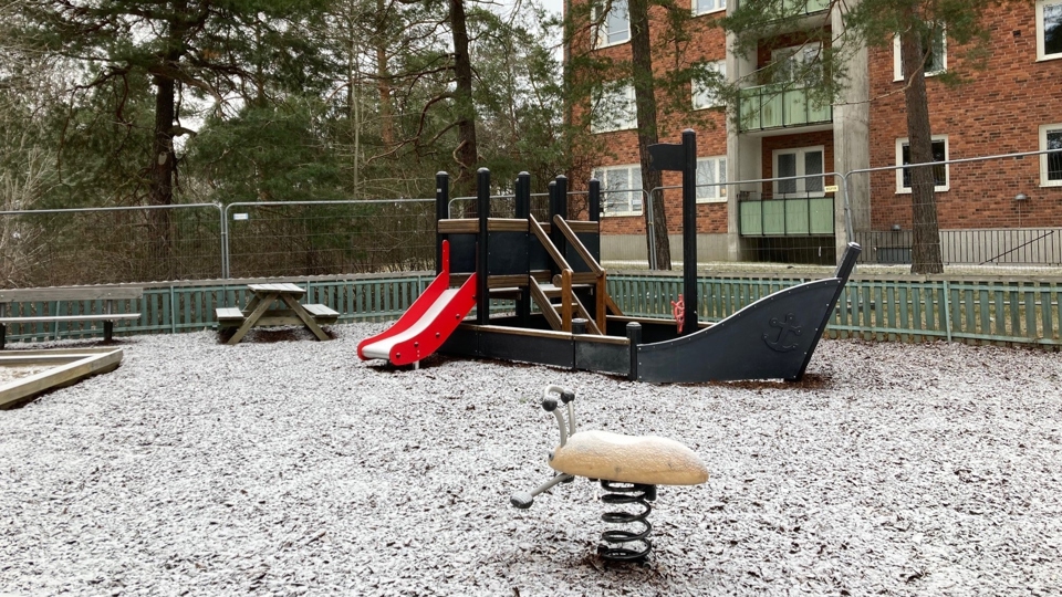 Lekplats omgärdad av staket med sandlåda, bänkar, gungdjur och en stor båt som man kan klättra i och åka rutschbana ifrån. I bakgrunden syns ett flerfamiljshus.