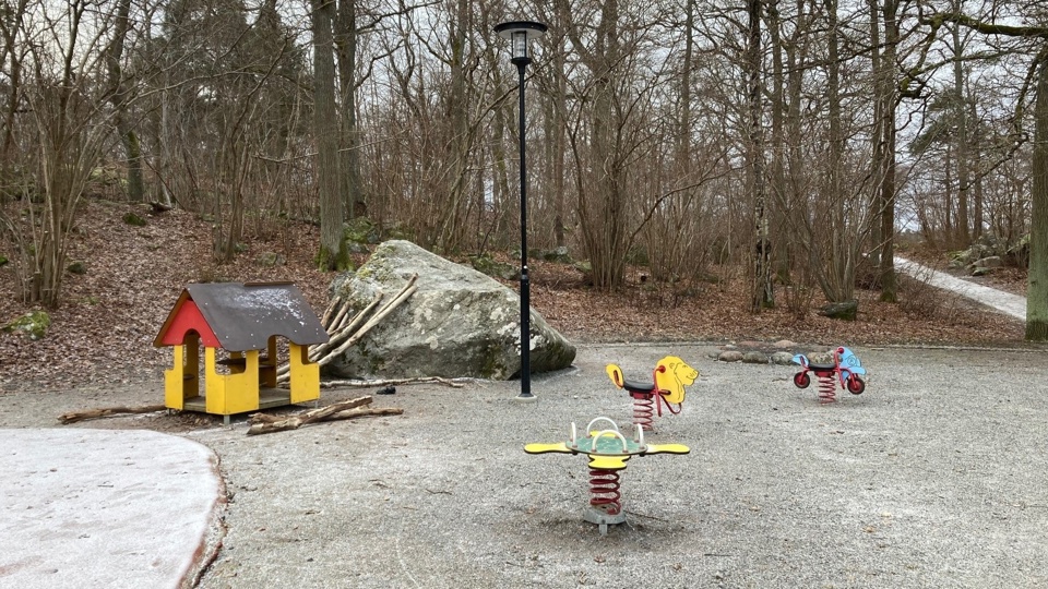 En lekstuga och flera gungsdjur vid en stor sten. Bredvid lekplatsen finns en skog med många stora stenar.