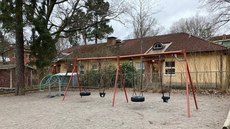 Yta med sand. Gungställning med fyra gungor på. I bakgrunden träd buskar och enplansfastighet. Foto.