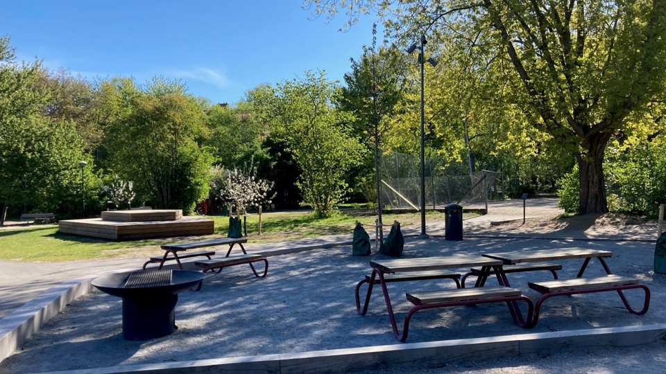 Picknickbord och grillplats. Träd och bollplan i bakgrunden. Foto. 