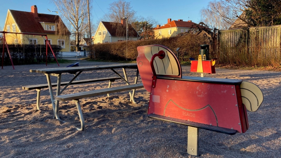 Lekplats inramad av buskar med picknickbord, gungdjur och gungor. Villor syns i bakgrunden.