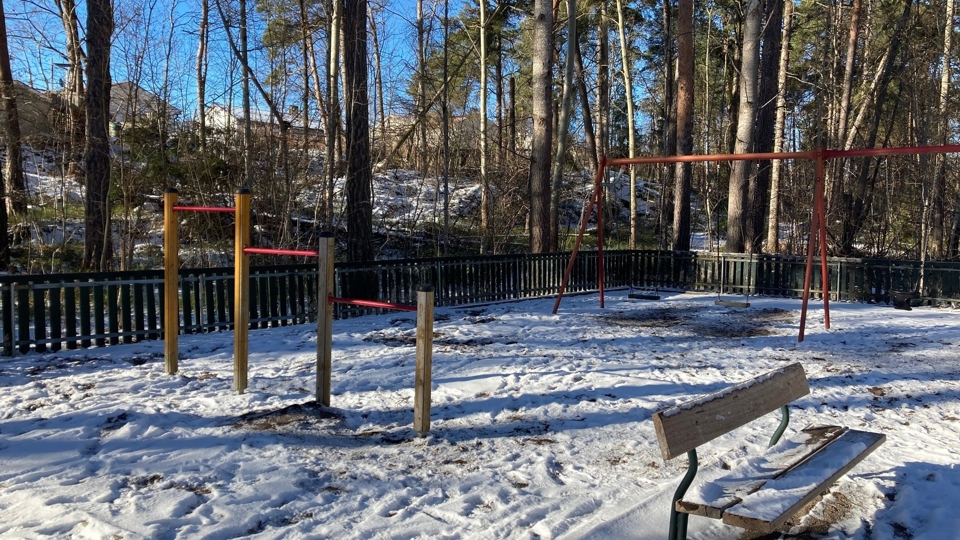 Lekplats med volträcke, bänk och gungor samt träd och flerfamiljshus i bakgrunden.