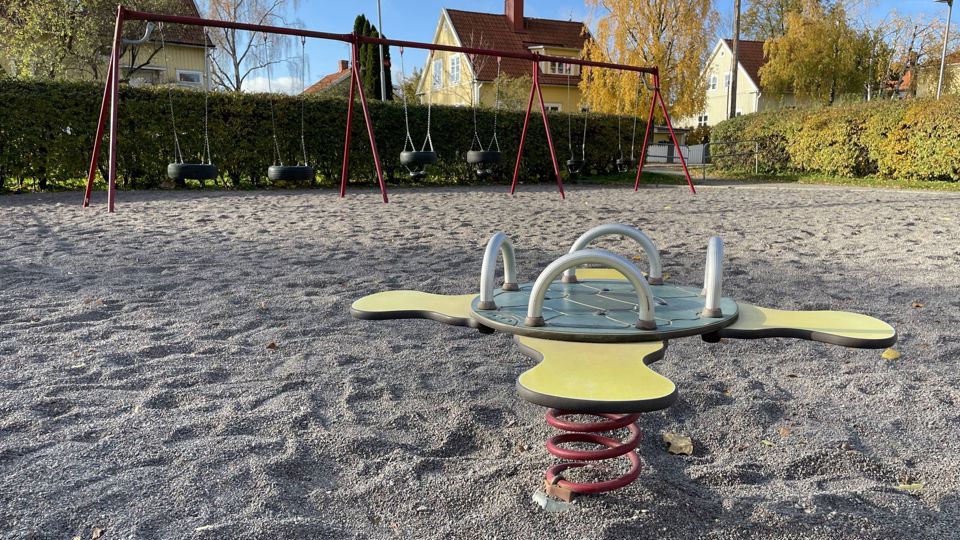 Lekplats inramad av buskar med många gungor. Villor syns i bakgrunden.