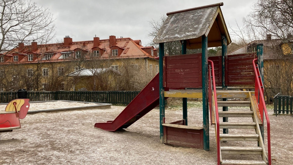 Lekplats med gunghästar och rutschkana med flerfamiljshus och träd i bakgrunden.