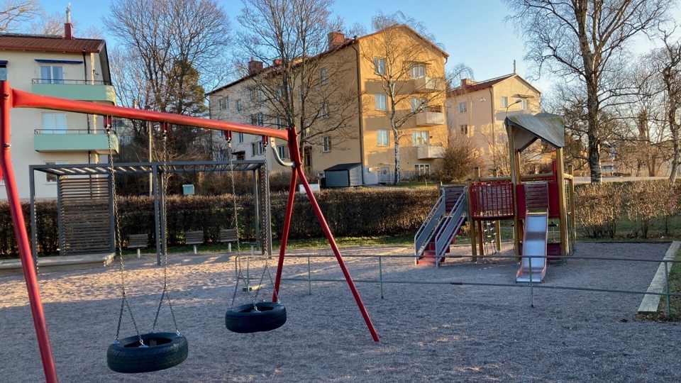 Lekplats inramad av buskar med gungor, klätterställning och pergola med sandlåda och sittplatser. Flerfamiljshus syns i bakgrunden.