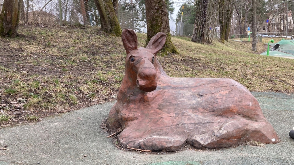 En skulptur av en liggande älg, i bakgrunden en slänt med höga träd. 