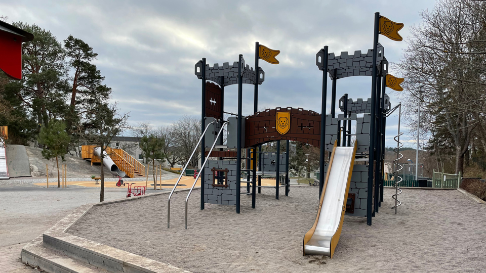 En borgliknande klätterställning med torn, flaggor och rutschkana som står i sanden.