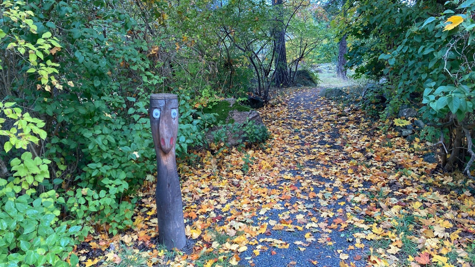 Ingång till lekplats, kantad av träfigur och lövad gångbana, foto.
