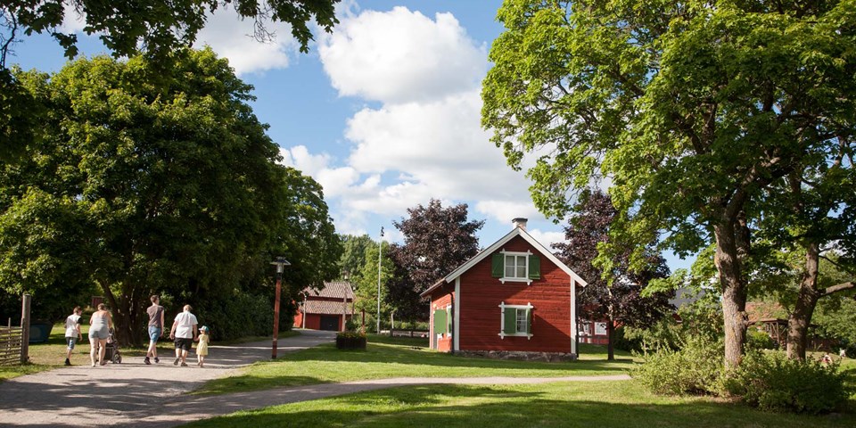 Ett gammalt rött hus med vita knutar och gröna fönsterluckor omgivet av stora gröna lövträd.  En familj går på grusvägen som går förbi huset.