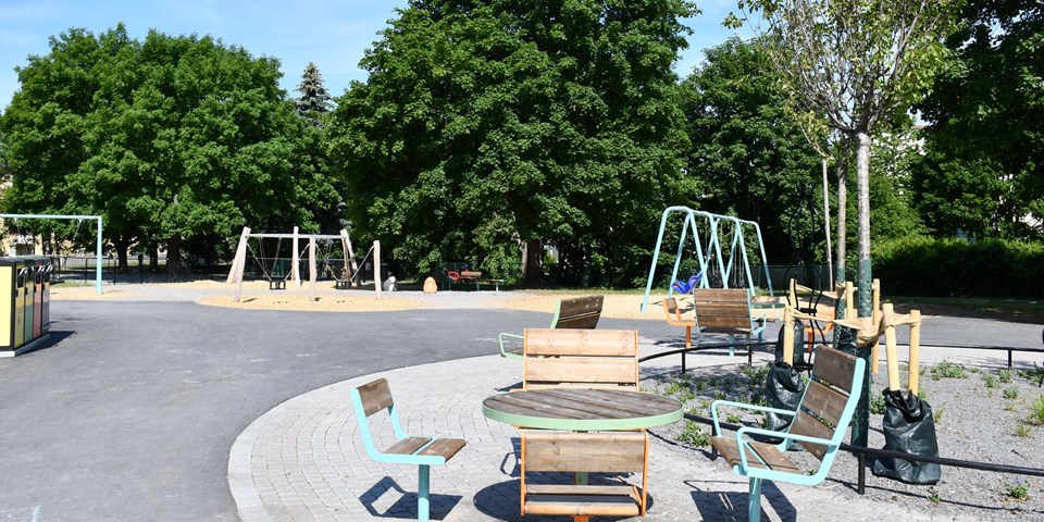 Park med gungor, stolar och bord, träd, buskar, gungor och källsorteringskärl. Foto.
