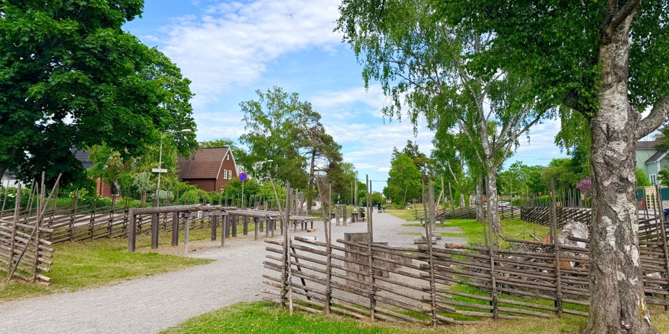 Grusgång genom ett grönområde med trästaket, klätterlek och stora lövträd på båda sidor.