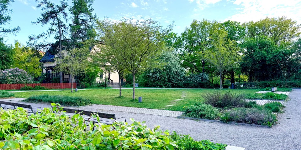 Grön park med gräsmatta, träd, planteringar och grusgångar. I bakgrunden syns en äldre byggnad omgiven av grönska.