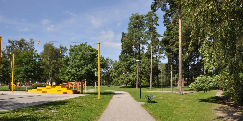 Gångväg genom Melonparken. Omgivande träd och gräs. Bänkar, lykstolpar och en lekplats. Foto.