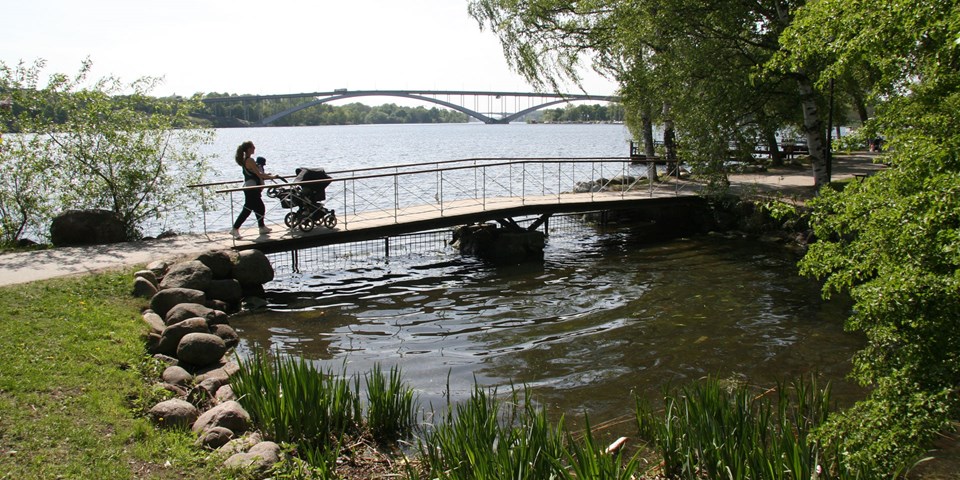 En kvinna med en barnvagn går över en bro på strandpromenaden vid Norr Mälarstrand.