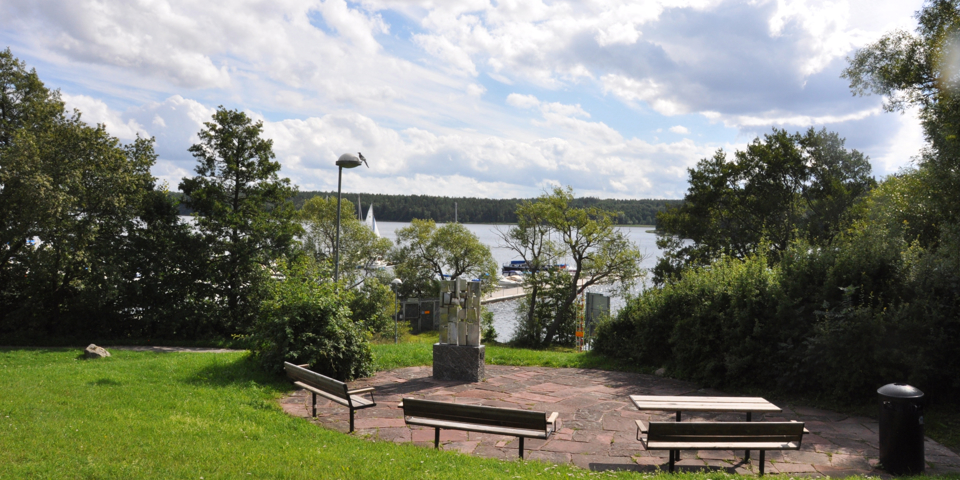 Bänkar och bord som är vända mot vattnet och båtbryggan. Bänkarna och bordet är placerade på en stenbeklädd plats som är omgiven av gräs, buskar, träd, konstverk, soptunna och lyktstolpe. Foto.