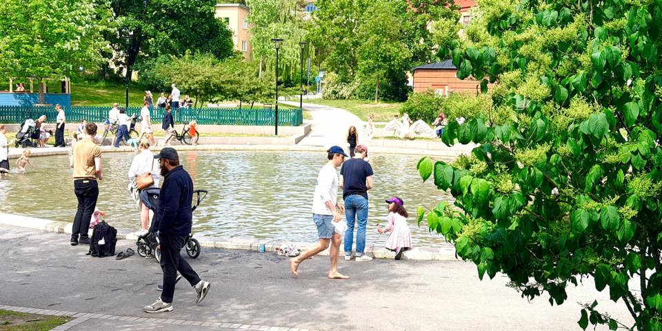 Besökare promenerar och vistas runt en damm i parken. Barn leker vid vattenkanten och grönskande träd omger området.
