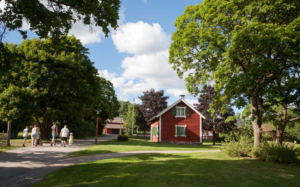 Ett gammalt rött hus med vita knutar och gröna fönsterluckor omgivet av stora gröna lövträd.  En familj går på grusvägen som går förbi huset.
