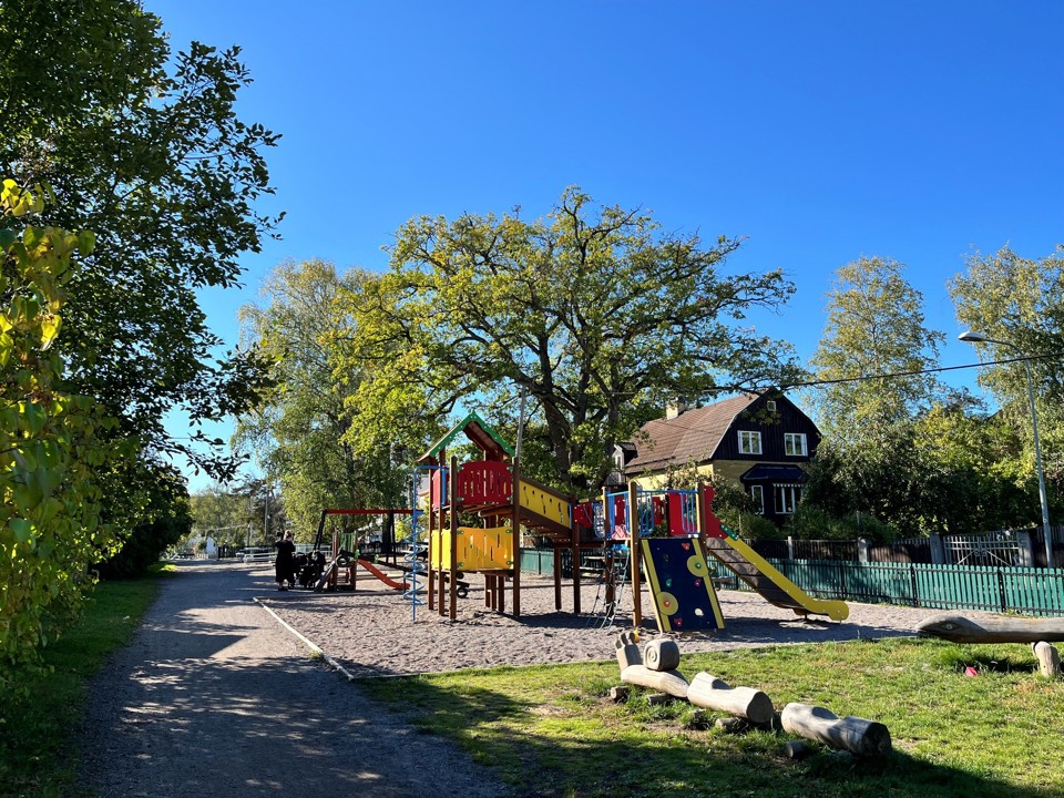 Lekplats med klätterställning och rutschkanor intill en grusgång med träd och villor i bakgrunden.