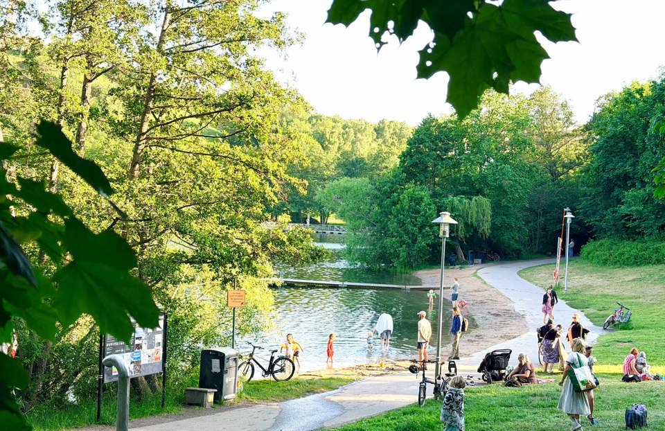 Människor badar och umgås vid Trekantsparkens badplats en solig sommardag. En liten sandstrand, brygga och gångväg omges av grönskande träd.