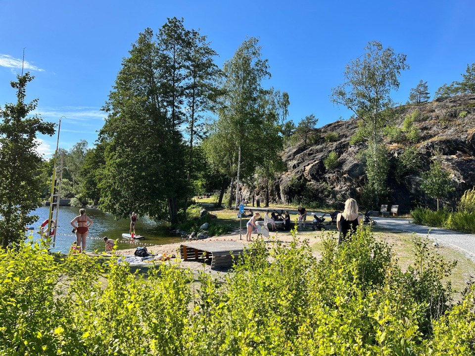 Badplats i Vinterviksparken där människor badar, sitter på stranden och umgås. Intill stranden finns klippor, träd och en gångväg.