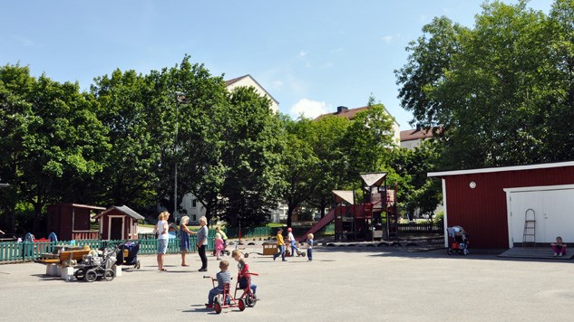 Lekplats med klätterställningar, lekredskap och en stor plan där barn cyklar på trehjulingar.