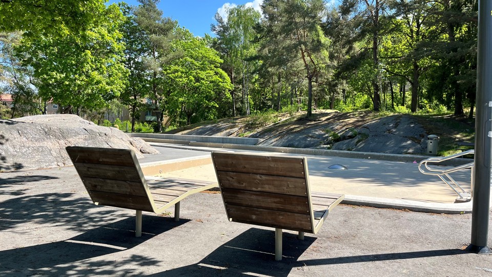 Två solsängar i trä framför en tömd plaskdamm i ett grönområde.