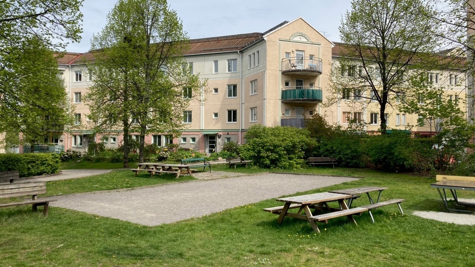 Picknickbord runt en grusad boulbana, ytan ramas in av träd med flerfamiljshus i bakgrunden.