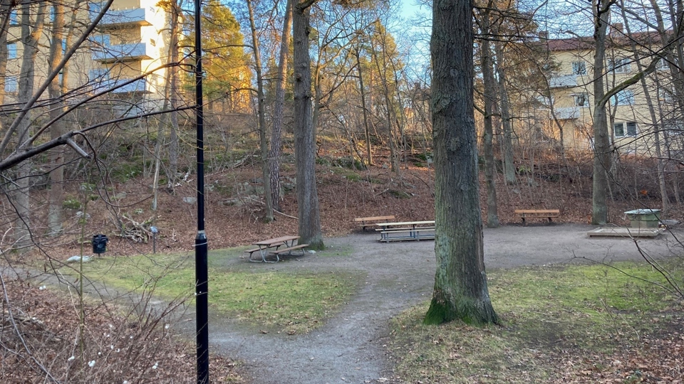 Lekplats med sandlåda och parkbänkar, omgiven av träd och med flerfamiljshus i bakgrunden. 