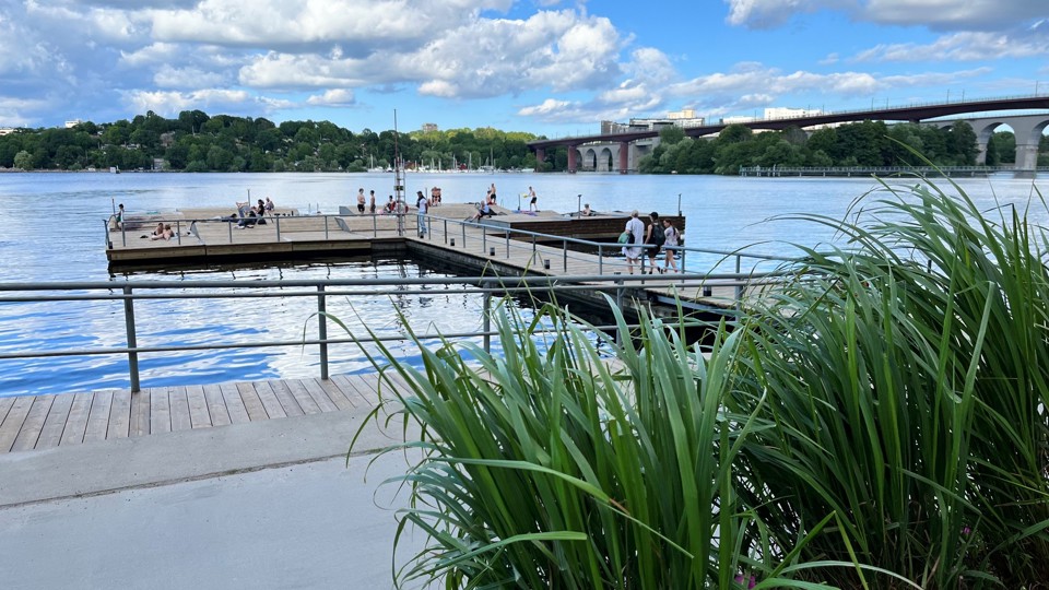 Ett promenadstråk längs vattnet och i vattnet ligger en stor bryggplats där människor solar och badar. Till höger syns en bro som sträcker sig till andra sidan vattnet.