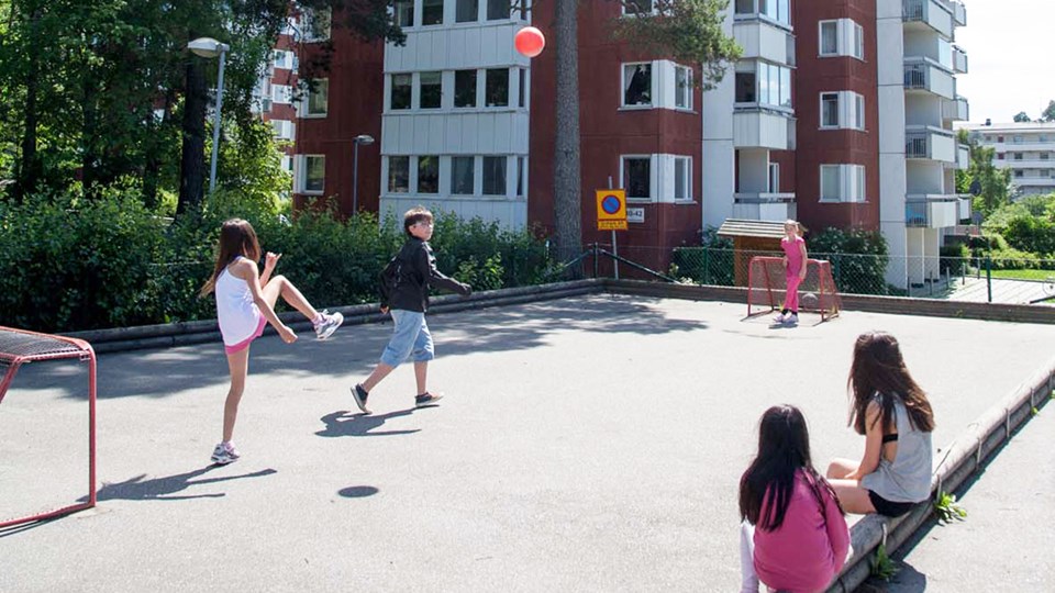 Fem barn spelar med röd boll på asfalterad plan med låg sarg. Två flickor sitter och tittar på.