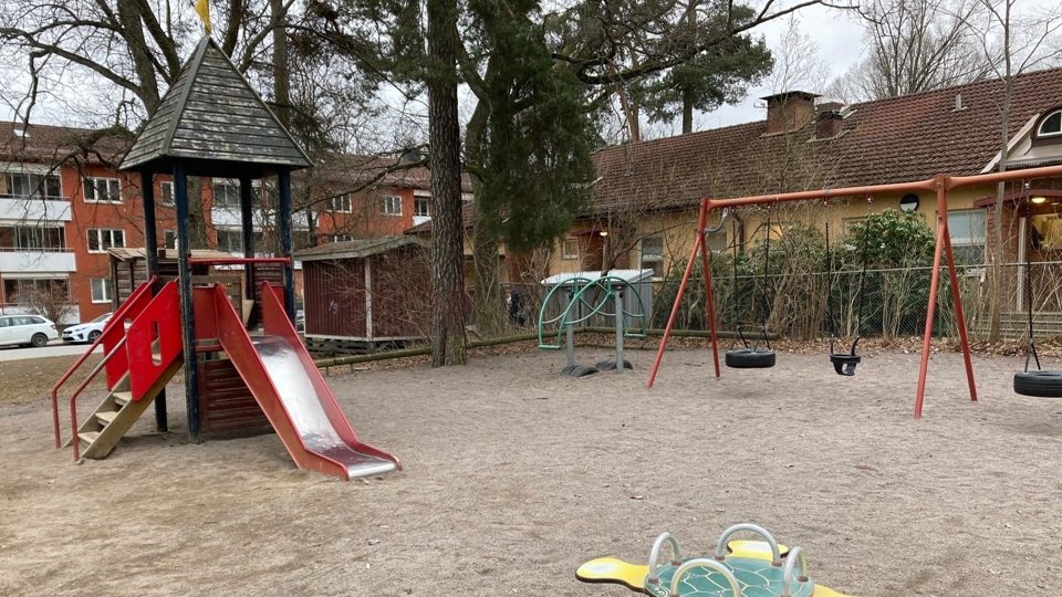 Stor yta med sand. Liten klätterrutchkana, vippgunga och gungställning. I bakgrunden finns flerfamiljshus, träd och buskar, foto