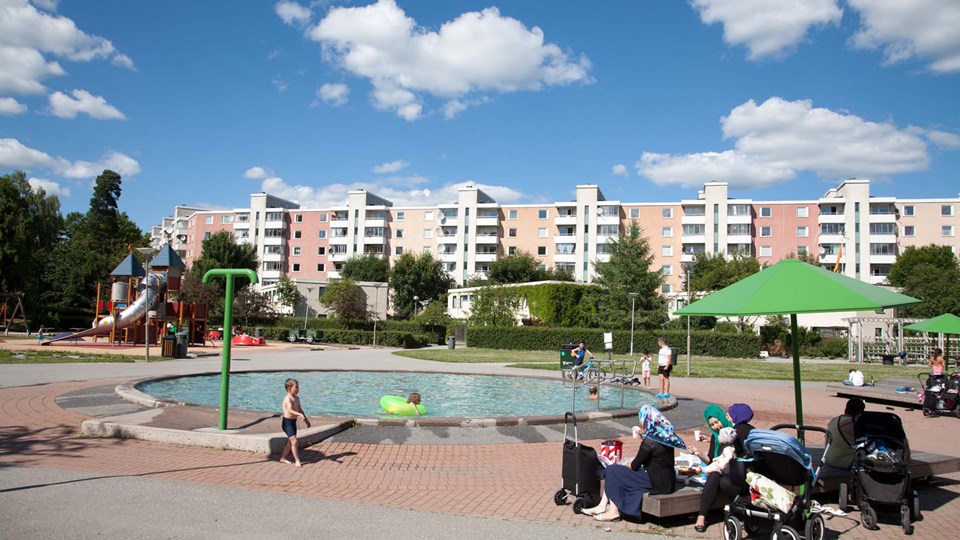 Öppen lekpark med plaskdamm och gröna parasoller i förgrunden och bostadshus i bakgrunden.