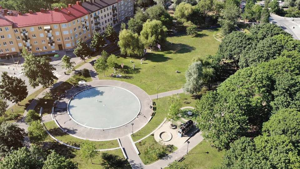 Flera gula flerfamiljshus med en rund plaskdamm i en grön park. Till plaskdammen finns flera gångvägar genom parken och de gröna gräsmattorna.