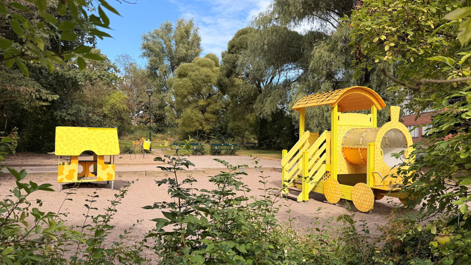 Lekplats i park med gul leksaksställning, sand och grönskande träd.
