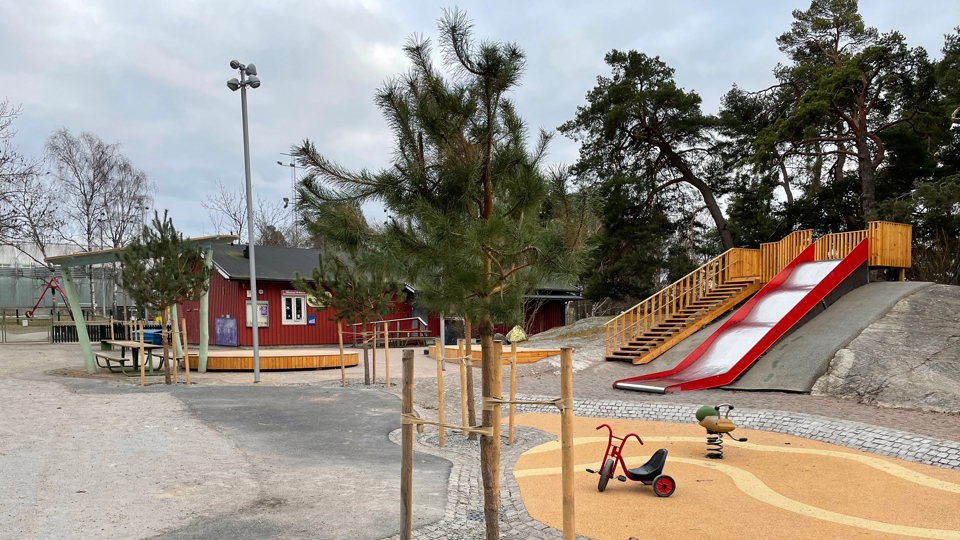 En lekplats med röd rutschkana i en backe, liten röd byggnad, träd och en trehjuling på marken.