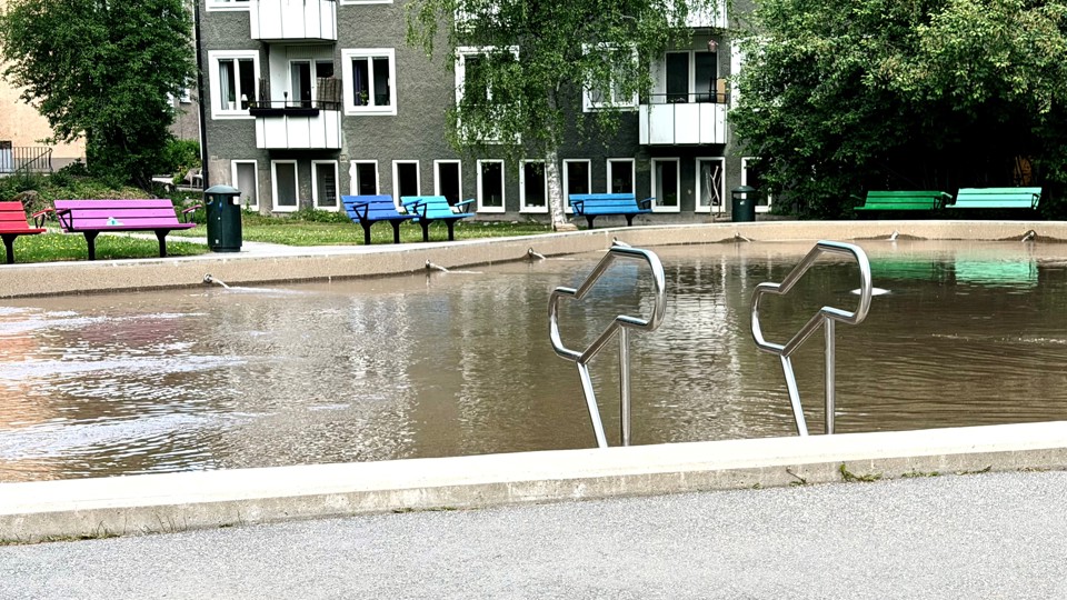 Plaskdamm framför ett grått flerfamiljshus. Längs kanten av plaskdammen finns färgglada parkbänkar i grönt, blått och rosa.