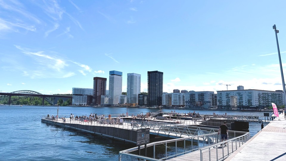 Stor bryggplats med badande och solbadande personer. På andra sidan vattnet syns flera höga flerfamiljshus i svart, vitt och rostfärgat.