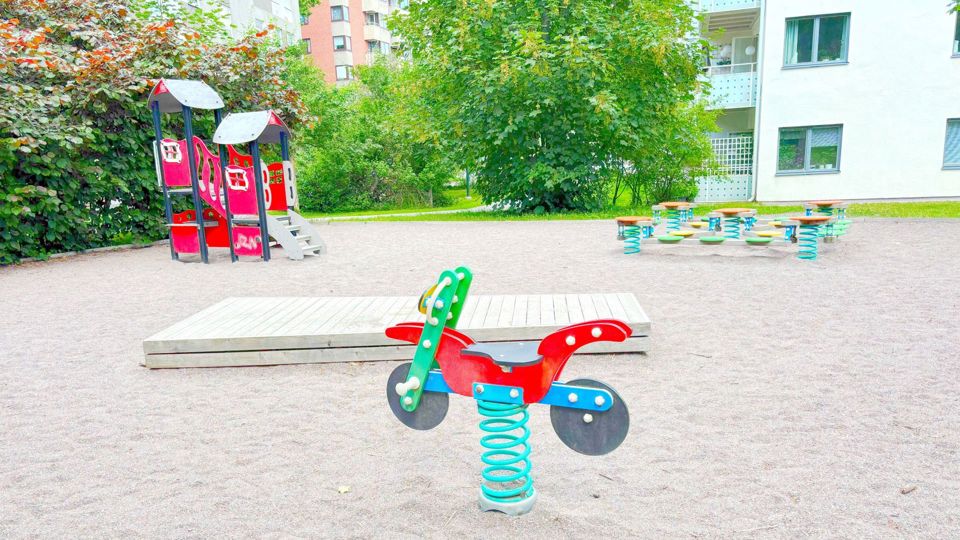 Lekplats med klätterborg, gungdjur och balanslek framför ett bostadshus.