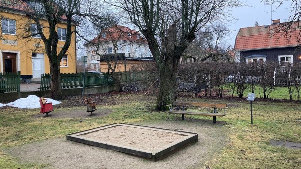 Lekplats med gungdjur, sandlåda och bänk. I bakgrunden syns flerfamiljshus och en villa.