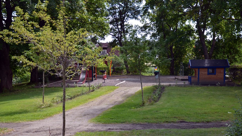 Lekparken i bakgrunden med lekstuga och rutschkana omgiven av träd.