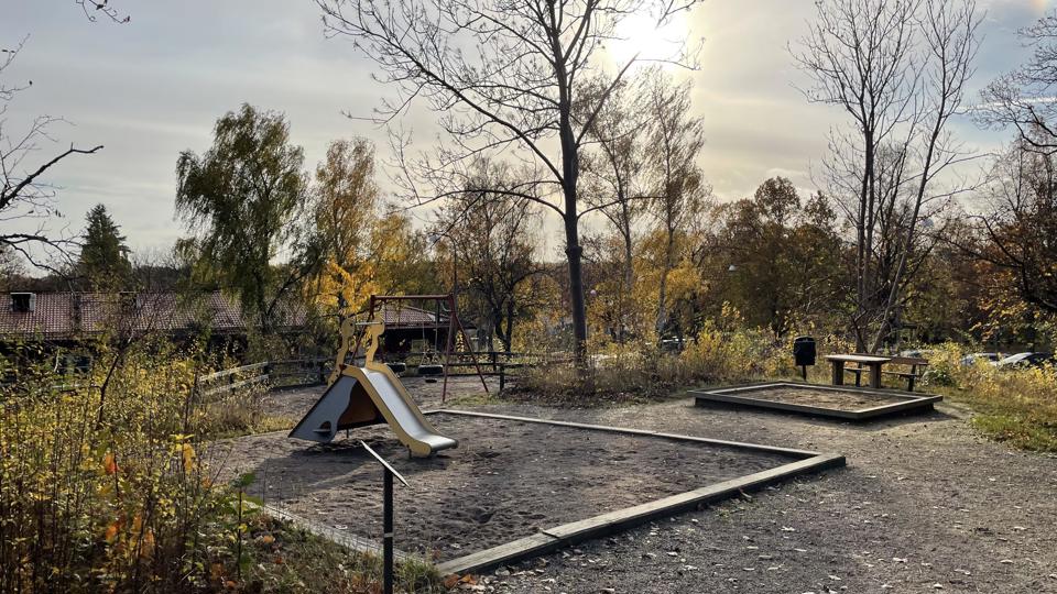 Lekplats omgärdad av träd med en förskola i bakgrunden. Lekplatsen är utrustad med rutschkana, gungor, sandlåda och en bänk med ett bord.