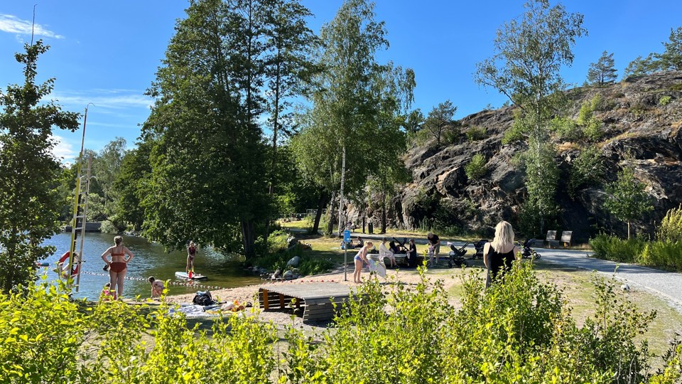 Solbadande badgäster på och i vattnet. En person paddlar på en stand up paddle board och vuxna torkar barn med handduk efter bad.